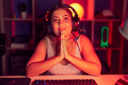 Photo for Young blonde woman playing video games wearing headphones praying with hands together asking for forgiveness smiling confident. - Royalty Free Image