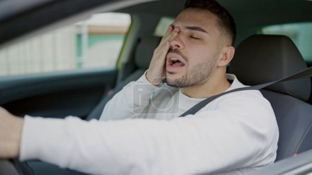 Foto de Joven hispano cansado conduciendo coche y bostezando en la calle - Imagen libre de derechos