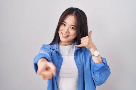 Foto de Joven mujer china de pie sobre fondo blanco sonriendo haciendo hablar por teléfono gesto y señalando a usted. Llámame.. - Imagen libre de derechos
