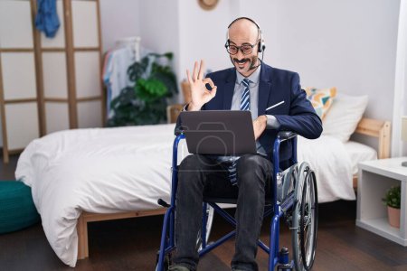 Téléchargez les photos : Homme hispanique avec barbe assis sur fauteuil roulant faire des affaires appel vidéo faire ok signe avec les doigts, sourire geste amical excellent symbole - en image libre de droit