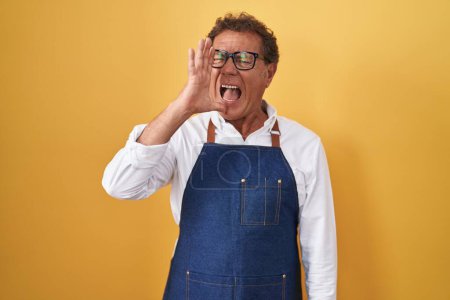 Foto de Hombre hispano de mediana edad con delantal de cocinero profesional gritando y gritando fuerte a un lado con la mano en la boca. concepto de comunicación. - Imagen libre de derechos