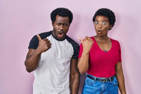 Téléchargez les photos : Jeune couple afro-américain debout sur fond rose surpris pointant du doigt sur le côté, bouche ouverte expression étonnée. - en image libre de droit