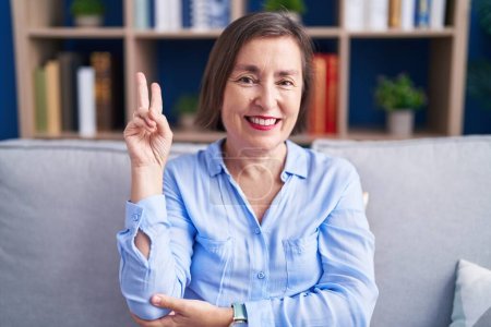 Photo for Middle age hispanic woman sitting on the sofa at home smiling with happy face winking at the camera doing victory sign. number two. - Royalty Free Image