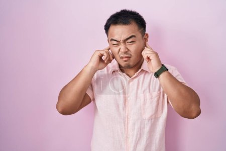 Foto de Joven chino de pie sobre fondo rosa cubriendo las orejas con los dedos con expresión molesta por el ruido de la música fuerte. concepto de sordo. - Imagen libre de derechos