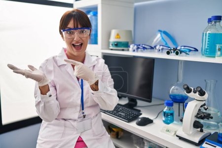 Téléchargez les photos : Jeune femme brune travaillant au laboratoire scientifique étonnée et souriante à la caméra tout en se présentant avec la main et pointant du doigt. - en image libre de droit