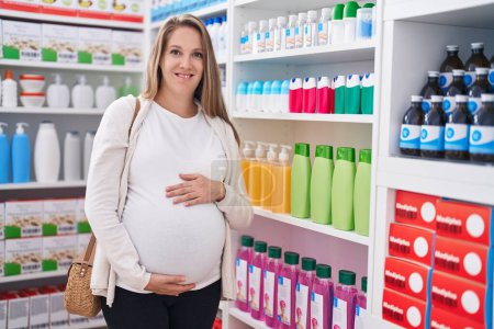 Téléchargez les photos : Young pregnant woman customer smiling confident touching belly at pharmacy - en image libre de droit
