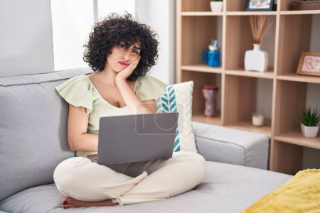 Téléchargez les photos : Jeune femme brune avec les cheveux bouclés à l'aide d'un ordinateur portable assis sur le canapé à la maison pensant fatigué et ennuyé avec des problèmes de dépression avec les bras croisés. - en image libre de droit