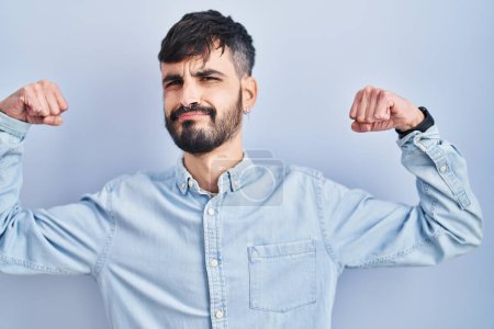 Téléchargez les photos : Jeune homme hispanique avec la barbe debout sur fond bleu montrant les muscles des bras souriant fier. concept de fitness. - en image libre de droit
