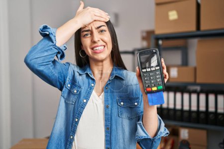 Photo for Hispanic woman working at small business ecommerce holding credit card and dataphone stressed and frustrated with hand on head, surprised and angry face - Royalty Free Image