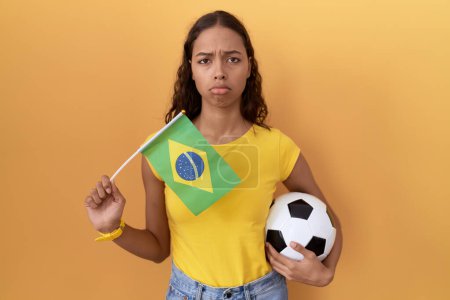 Foto de Mujer hispana joven sosteniendo bandera brasileña y pelota de fútbol deprimida y preocupada por la angustia, llorando enojada y asustada. expresión triste. - Imagen libre de derechos