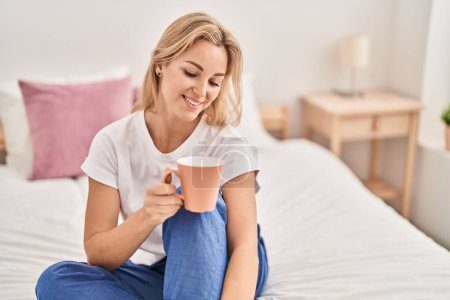 Foto de Mujer rubia joven bebiendo taza de café sentado en la cama en el dormitorio - Imagen libre de derechos