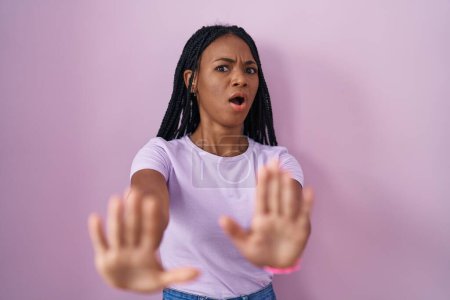 Foto de Mujer afroamericana con trenzas de pie sobre fondo rosa asustada y aterrorizada con la expresión del miedo detienen el gesto con las manos, gritando en shock. concepto de pánico. - Imagen libre de derechos