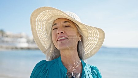 Foto de Mujer turista de pelo gris de mediana edad sonriendo confiada de pie en la playa - Imagen libre de derechos