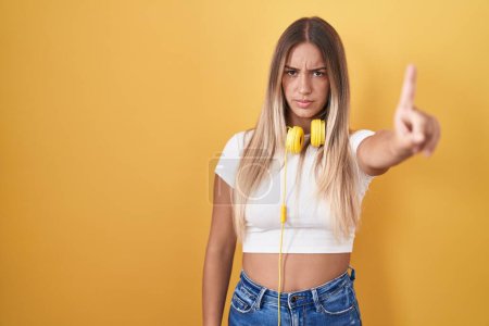 Téléchargez les photos : Jeune femme blonde debout sur fond jaune portant des écouteurs pointant du doigt et l'expression en colère, ne montrant aucun geste - en image libre de droit