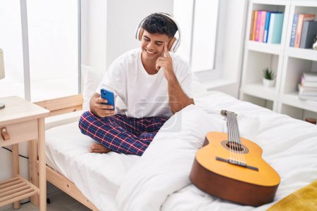 Foto de Joven hispano escuchando música sentado en la cama en el dormitorio - Imagen libre de derechos