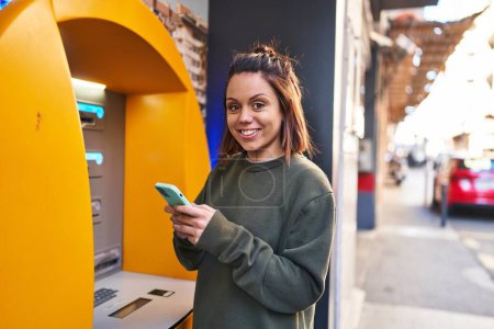Photo for Young beautiful hispanic woman using smartphone standing by bank teller at street - Royalty Free Image
