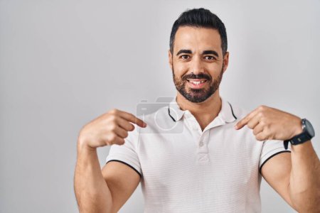 Foto de Joven hombre hispano con barba vistiendo ropa casual sobre fondo blanco mirando confiado con sonrisa en la cara, señalándose con los dedos orgullosos y felices. - Imagen libre de derechos