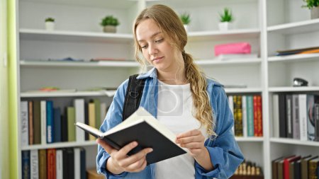 Photo for Young blonde woman student standing reading book at library university - Royalty Free Image