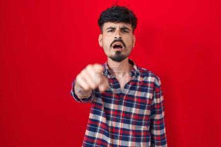 Téléchargez les photos : Jeune homme hispanique avec la barbe debout sur fond rouge pointant mécontent et frustré à la caméra, en colère et furieux contre vous - en image libre de droit