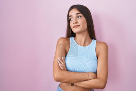 Téléchargez les photos : Jeune femme brune debout sur fond rose regardant sur le côté avec les bras croisés convaincus et confiants - en image libre de droit