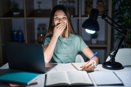 Foto de Adolescente chica haciendo la tarea en casa tarde en la noche riendo y riéndose avergonzado cubriendo la boca con las manos, chismes y concepto de escándalo - Imagen libre de derechos