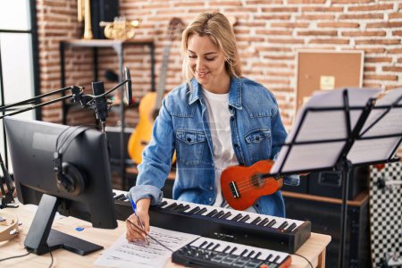Photo for Young blonde woman musician composing song holding ukelele at music studio - Royalty Free Image