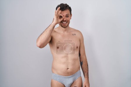 Téléchargez les photos : Jeune homme hispanique debout torse nu portant des sous-vêtements faisant un geste correct avec la main souriante, les yeux regardant à travers les doigts avec un visage heureux. - en image libre de droit