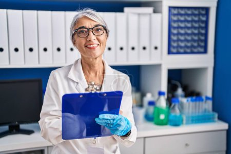 Téléchargez les photos : Moyen âge femme aux cheveux gris scientifique souriant confiant écrire sur presse-papiers au laboratoire - en image libre de droit