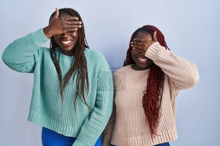Photo for Two african woman standing over blue background smiling and laughing with hand on face covering eyes for surprise. blind concept. - Royalty Free Image