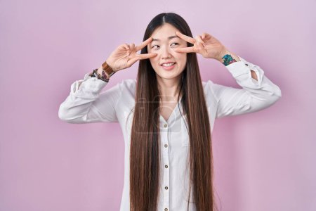 Téléchargez les photos : Chinese young woman standing over pink background doing peace symbol with fingers over face, smiling cheerful showing victory - en image libre de droit