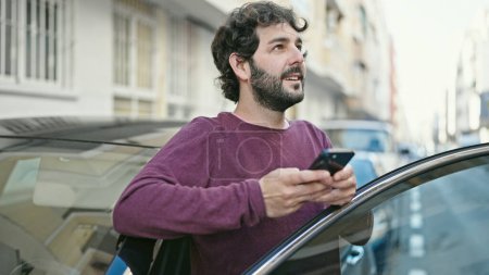 Foto de Young hispanic man using smartphone leaning on car at street - Imagen libre de derechos