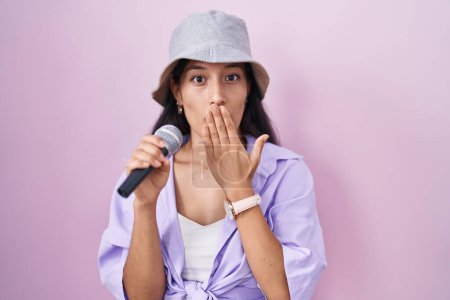 Téléchargez les photos : Jeune femme hispanique chantant chanson à l'aide d'un microphone sur fond rose couvrant la bouche avec la main, choquée et effrayée par erreur. expression surprise - en image libre de droit