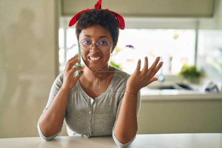 Téléchargez les photos : Femme afro-américaine parlant sur smartphone assis sur la table à la maison - en image libre de droit