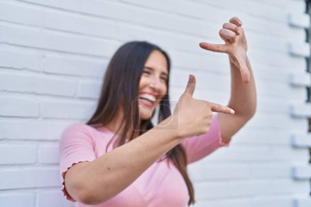 Foto de Joven hermosa mujer hispana sonriendo confiada haciendo gesto fotográfico con las manos sobre el fondo de ladrillo blanco aislado - Imagen libre de derechos