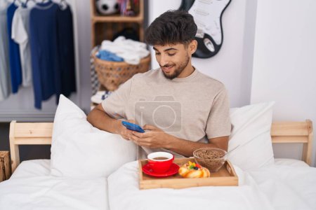 Foto de Hombre árabe joven usando teléfono inteligente desayunando en el dormitorio - Imagen libre de derechos