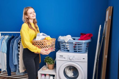 Foto de Joven mujer rubia sonriendo confiado celebración cesta con ropa en la sala de lavandería - Imagen libre de derechos