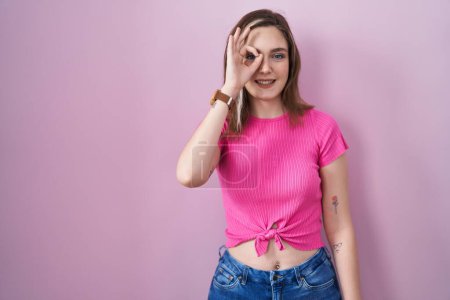 Photo for Blonde caucasian woman standing over pink background doing ok gesture with hand smiling, eye looking through fingers with happy face. - Royalty Free Image