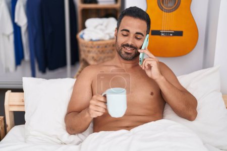 Foto de Joven hispano hablando en smartphone bebiendo café tumbado en la cama en el dormitorio - Imagen libre de derechos