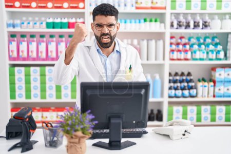 Photo for Hispanic man with beard working at pharmacy drugstore angry and mad raising fist frustrated and furious while shouting with anger. rage and aggressive concept. - Royalty Free Image