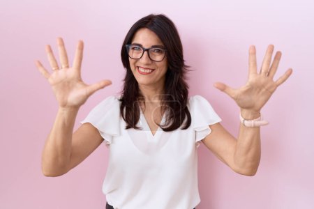Téléchargez les photos : Femme hispanique du Moyen Âge portant un t-shirt blanc décontracté et des lunettes montrant et pointant vers le haut avec les doigts numéro dix tout en souriant confiant et heureux. - en image libre de droit