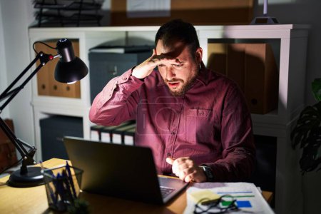 Photo for Plus size hispanic man with beard working at the office at night very happy and smiling looking far away with hand over head. searching concept. - Royalty Free Image