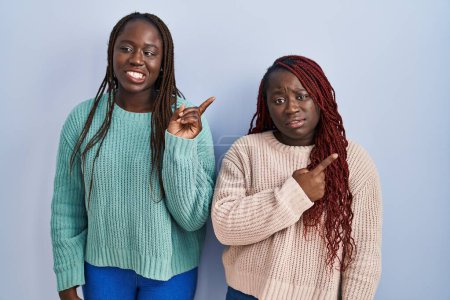 Foto de Dos mujeres africanas de pie sobre fondo azul señalando a un lado preocupado y nervioso con el dedo índice, preocupado y expresión sorprendida - Imagen libre de derechos