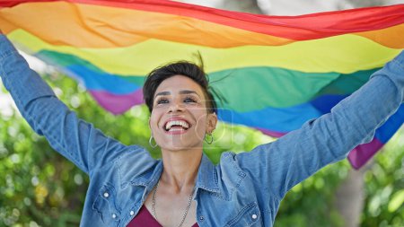 Foto de Joven hermosa mujer hispana sonriendo confiada sosteniendo la bandera del arco iris en el parque - Imagen libre de derechos