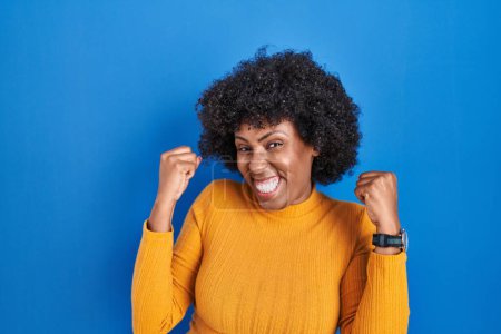 Téléchargez les photos : Femme noire avec les cheveux bouclés debout sur fond bleu célébrant surpris et étonnés pour le succès avec les bras levés et les yeux fermés. concept gagnant. - en image libre de droit
