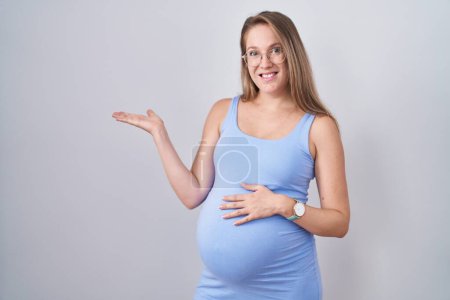 Foto de Mujer embarazada joven de pie sobre fondo blanco señalando a un lado con las manos abiertas palmeras mostrando espacio de copia, presentando anuncio sonriente emocionado feliz - Imagen libre de derechos