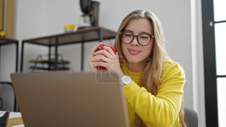 Foto de Mujer joven rubia trabajadora de negocios usando laptop bebiendo café en la oficina - Imagen libre de derechos