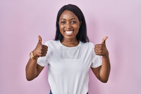 Foto de Mujer joven africana vistiendo casual camiseta blanca signo de éxito haciendo gesto positivo con la mano, pulgares hacia arriba sonriendo y feliz. expresión alegre y gesto ganador. - Imagen libre de derechos