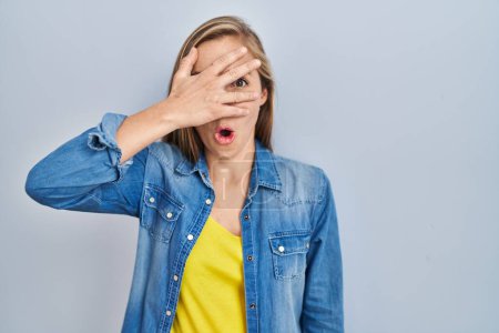 Photo for Young blonde woman standing over blue background peeking in shock covering face and eyes with hand, looking through fingers with embarrassed expression. - Royalty Free Image