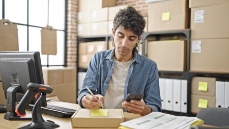 Téléchargez les photos : Jeune homme hispanique ecommerce travailleur d'affaires en utilisant l'écriture smartphone sur le paquet au bureau - en image libre de droit
