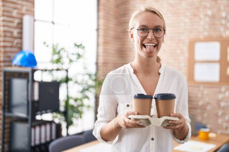 Téléchargez les photos : Jeune femme caucasienne travaillant au bureau tenant des tasses à café collant la langue heureux avec une expression drôle. - en image libre de droit